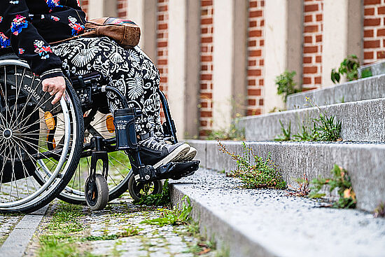 Zu sehen ist eine Person im Rollstuhl. Sie steht mit dem Rollstuhl vor einer Treppe. Die Treppe stellt eine Barriere dar. Zu sehen ist eine Person im Rollstuhl. Sie steht mit dem Rollstuhl vor einer Treppe. Die Treppe stellt eine Barriere dar.